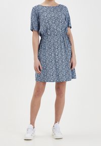 Osoba v modro-bílé květinové šatech s krátkým rukávem a délky ke kolenům, obutá v bílé sportovní tenisky, stojící na jednobarevném pozadí.