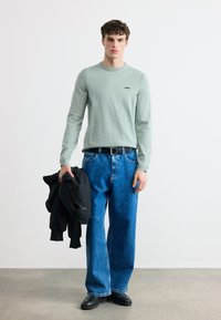 Νεαρός άνδρας που φορά ανοιχτό πράσινο μακρυμάνικο πουκάμισο, μπλε τζιν, μαύρη ζώνη και παπούτσια, κρατώντας μαύρο μπουφάν στο δεξί χέρι, στέκεται σε γκρι πάτωμα.