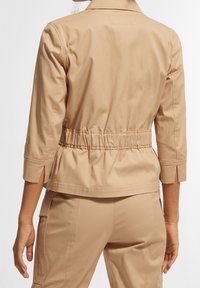 Beżowa kurtka typu cropped z dopasowaną talią, wyrazistym kołnierzem i rękawami o długości trzech kwart. Gładka tekstura z bocznymi rozcięciami i subtelnymi detalami stębnowania.