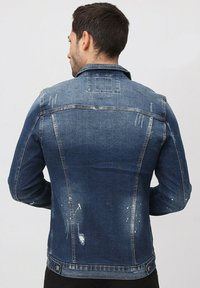 Veste en denim bleu marine avec une finition usée, présentant des motifs d'usure et un léger délavage. Comprend un col classique et une fermeture à boutons.