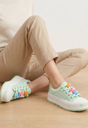 Person, die beigen Hosen trägt, sitzt auf dem Holzboden und zeigt hellgrüne Plateausneaker mit bunten Kunststoffanhängern an den Schnürsenkeln.