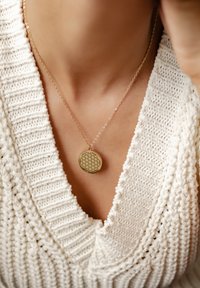 Collar de oro con un patrón geométrico, reposando sobre un suéter blanco texturizado, con un diseño de escote en V amplio.