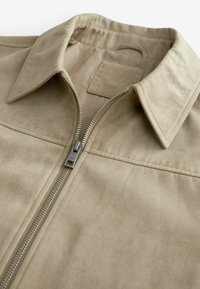 Veste en suédine beige avec un col accentué, fermeture à glissière sur le devant, couture décorative et une finition douce et texturée.
