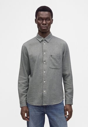 Homme portant une chemise grise à manches longues avec une poche poitrine et un jean bleu, debout devant un fond uni.