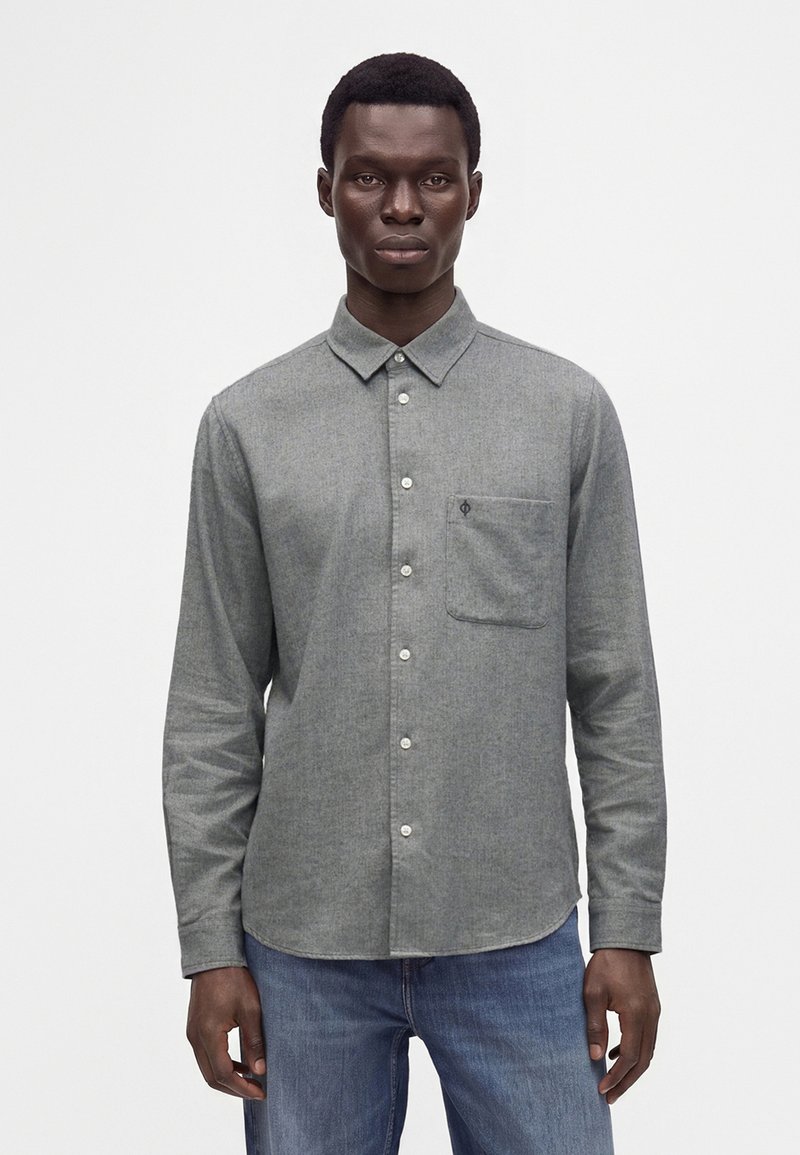 Homme portant une chemise grise à manches longues avec une poche poitrine et un jean bleu, debout devant un fond uni.