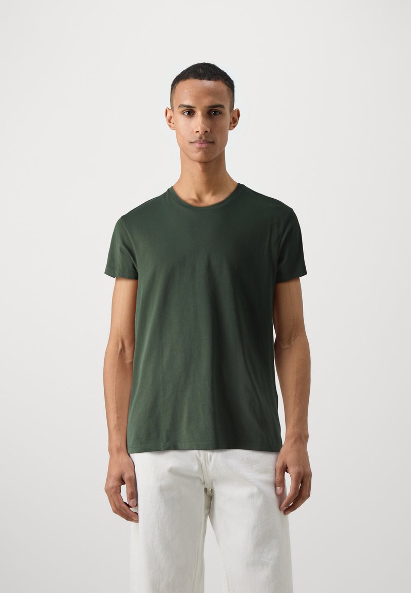 Grünes Rundhals-T-Shirt aus weichem Baumwollstoff. Kurze Ärmel und eine lockere Passform, kombiniert mit weißen geraden Hosen. Minimales Design.