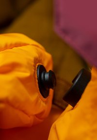 Tessuto arancione brillante con una texture liscia, caratterizzato da una manopola nera di regolazione e cuciture a contrasto. Primo piano dei dettagli del capo.