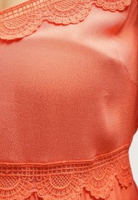 Korallfärgad topp med en texturerad tyg och spetskant längs halsringningen, som visar upp intrikata vågiga mönster och en mjuk glans.