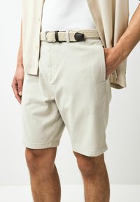 Pantaloni scurți bej deschis cu un finisaj texturat, având buzunare laterale și o curea împletită cu o cataramă închisă. Stilizați cu o cămașă deschisă.