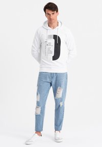 Joven con una sudadera blanca estampada, jeans claros rasgados y zapatillas blancas, de pie con las manos detrás de la espalda sobre un fondo blanco.