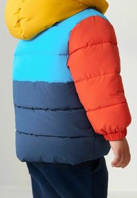 Giacca a vento color block con cappuccio giallo, che presenta sezioni blu, rosse e blu scuro. Il materiale appare trapuntato e isolato.