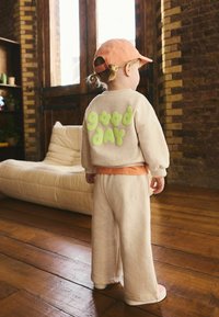 Sudadera de algodón beige con la inscripción "buen día" en letras verdes brillantes, combinada con pantalones beige a juego, y una camiseta interior y gorra naranjas.