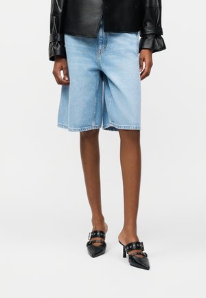 Személy világoskék farmer térd hosszúságú rövidnadrágot, fekete bőr dzsekit és fekete, csatos pántos, hegyes orrú, magas sarkú mules cipőt visel.