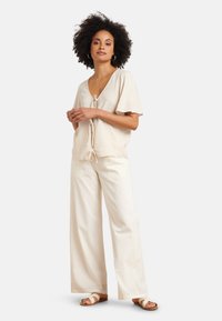 Helle beige Bluse mit kurzen, flatternden Ärmeln und einer Schleife vorne, kombiniert mit cremefarbenen, gestreiften, weiten Hosen und schlichten Sandalen.