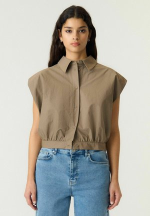 Mujer con cabello largo y oscuro lleva una blusa sin mangas color topo con botones y hombros anchos, y vaqueros azules de tiro alto sobre un fondo claro y liso.