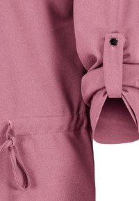 Vêtement rose à manches longues avec un revers roulé maintenu par une patte à bouton noir et un détail cordon près de l'ourlet.