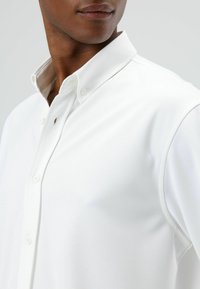 Weißes Hemd mit Knopfleiste und umgeschlagenem Kragen, das eine glatte Textur, klassische Passform und ein dezentes Logo-Detail in der Nähe der Knopfleiste aufweist.