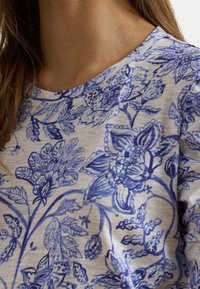 Kvinnans överkropp bär en beige skjorta med ett intrikat blått blommönster och blad, med långt brunt hår synligt till vänster.