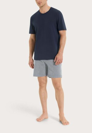 Uomo con indosso una maglietta blu navy a maniche corte e pantaloncini bianchi con sottili strisce verticali nere, in piedi a piedi nudi su uno sfondo chiaro.