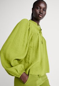 Blusa leggera verde lime con maniche voluminose, polsini arricciati e chiusura anteriore a nodo. Tessuto strutturato con una vestibilità comoda.