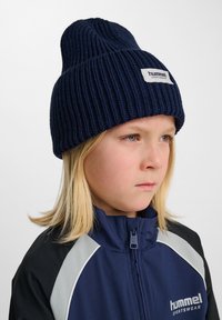 Marinblå ribbad stickad mössa med en vävd etikett med texten "hummel SPORTSWEAR." Personen bär en marinblå och svart jacka med ljusgrå accenter.