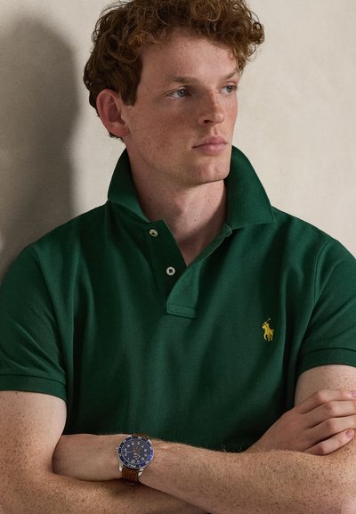 Νεαρός άνδρας με σγουρά κόκκινα μαλλιά φοράει σκούρο πράσινο πόλο με κίτρινο λογότυπο, καφέ δερμάτινο ρολόι, κοιτάζει πλάγια με σταυρωμένα τα χέρια.