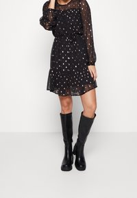 Ein Modell steht in einem schwarzen, langärmeligen Kleid mit einem dezenten Muster und einem ausgestellten Saum, kombiniert mit kniehohen schwarzen Stiefeln.