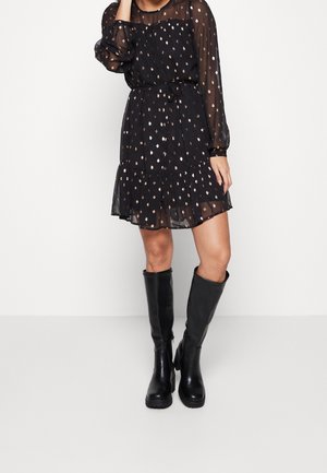 Ein Modell steht in einem schwarzen, langärmeligen Kleid mit einem dezenten Muster und einem ausgestellten Saum, kombiniert mit kniehohen schwarzen Stiefeln.