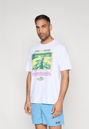 Biała bawełniana koszulka z kolorowym wzorem graficznym w zielone, żółte i różowe elementy. Model nosi jasnoniebieskie spodenki.