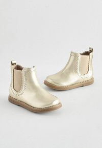 Stivali alla caviglia metallici in oro con una texture liscia, pannelli elastici beige a contrasto sui lati e un bordo decorativo a scallop sull'orlo.