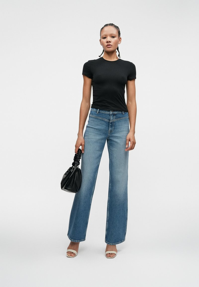 Czarna dopasowana koszulka, jasnoniebieskie szerokie dżinsy, przezroczyste sandały na obcasie oraz mała czarna torebka z marszczonym uchwytem. Gładkie białe tło.