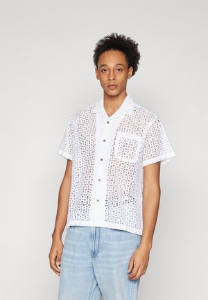 Short-sleeve, white shirt with a textured, open pattern, featuring a collared neckline and front pocket, paired with light blue jeans.