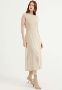 Beiges knielanges Kleid mit taillierter Silhouette, kurzen Ärmeln, seitlichem Schlitz und glattem Stoff. Kombiniert mit beigen, hochhackigen Stiefeletten.