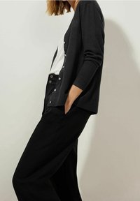 Mörkgrå cardigan med långa ärmar, framknäppning, bärs över en vit topp och kombineras med högmidjade svarta byxor. Slät textur.