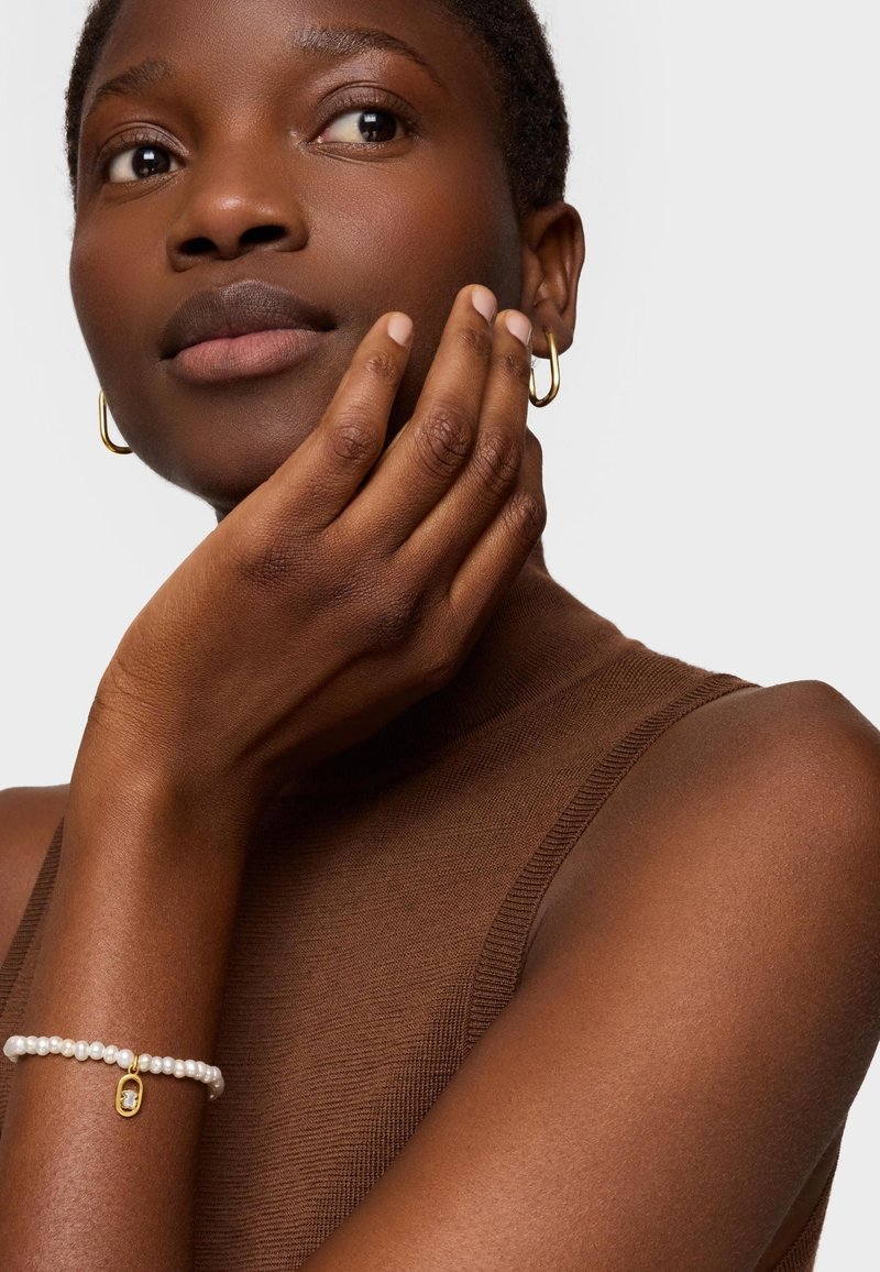 Mujer de cabello corto con aretes de aro dorados y una pulsera de perlas con un dije dorado, tocándose la mejilla, vestida con una blusa sin mangas color marrón.