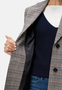Bruin en grijs houndstooth-gepatrooneerde jas met een gekartelde kraag, zwarte knopen en een gladde voering. Gelaagd over een marineblauw gebreid shirt.