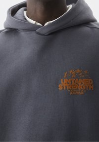 Grijze katoenen hoodie met een kangoeroezak, een capuchon met trekkoord en een oranje geborduurd logo met de tekst "UNTAMED STRENGTH" op de borst.