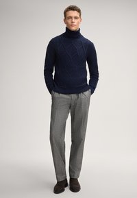 Uomo in piedi con le mani in tasca, che indossa un dolcevita a trecce di colore blu navy, pantaloni a quadri grigi e stivali scamosciati marroni scuri.
