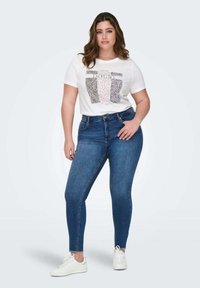 T-shirt blanc avec imprimé graphique en rose et noir, associé à un jean skinny bleu foncé. Le matériau semble doux avec une texture lisse. Baskets blanches.