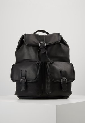 Sac à dos en cuir noir avec rabat supérieur, fermeture par cordon, deux poches avant à boucles et une poignée de transport sur un piédestal blanc.