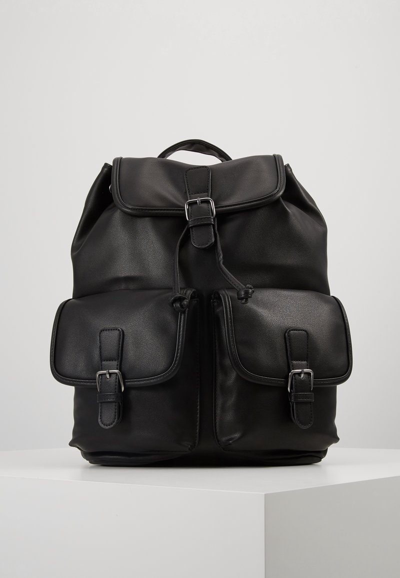 Sac à dos en cuir noir avec rabat supérieur, fermeture par cordon, deux poches avant à boucles et une poignée de transport sur un piédestal blanc.