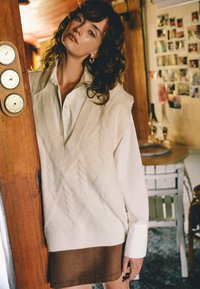 Gilet en tricot torsadé crème sur une chemise blanche à col, associé à une jupe marron. Cadre naturel avec des éléments en bois en arrière-plan.