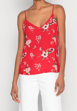 Femme portant un top rouge fleuri sans manches à fines bretelles et un pantalon blanc, debout sur un fond uni.