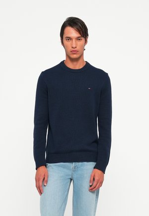 Man stands facing forward wearing a navy blue knit sweater and light blue jeans against a plain white background.