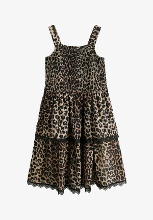 Dekle z dolgimi rjavimi lasmi, oblečeno v oprijeto obleko z leopardjim vzorcem brez rokavov in črnimi sandali z zaponko, stoji pred svetlo sivo ozadje.