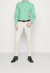 Zelená košile s kostkovaným vzorem na knoflíky, bílé kalhoty, černý pásek a černé šněrovací boty; hladká látka s přiléhavým střihem a upravenou siluetou.