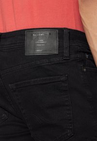 Vaqueros de denim negro con una etiqueta de cuero en la cinturilla. La etiqueta presenta texto estampado. La costura de los bolsillos es sutil y oscura.