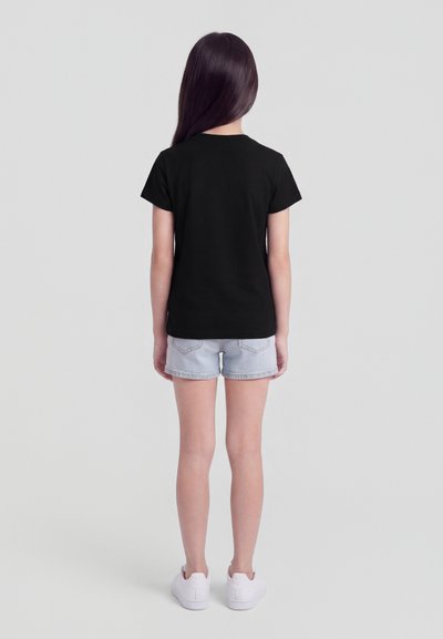 Παιδί με μακριά σκούρα μαλλιά, που φοράει απλό μαύρο μπλουζάκι, ανοιχτόχρωμο τζιν σορτσάκι και λευκά αθλητικά παπούτσια, στέκεται γυρισμένο προς τα πίσω πάνω σε γκρι φόντο.