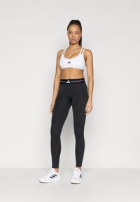 Juodos leginsės su aukštu liemeniu ir baltais prekės ženklo elementais, derinamos su baltu sportiniu liemenėliu, kuriame yra juodi akcentai ir plonos petnešėlės. Balti sportbačiai.