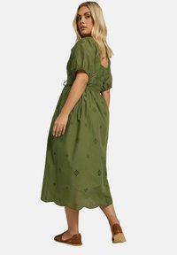 Vihreä puuvillainen midi-mekko, jossa on puffihihat, selässä leikkaus ja kirjailtu timanttikuvio; aaltoileva helma; yhdistetty ruskeisiin litteisiin sandaaleihin.